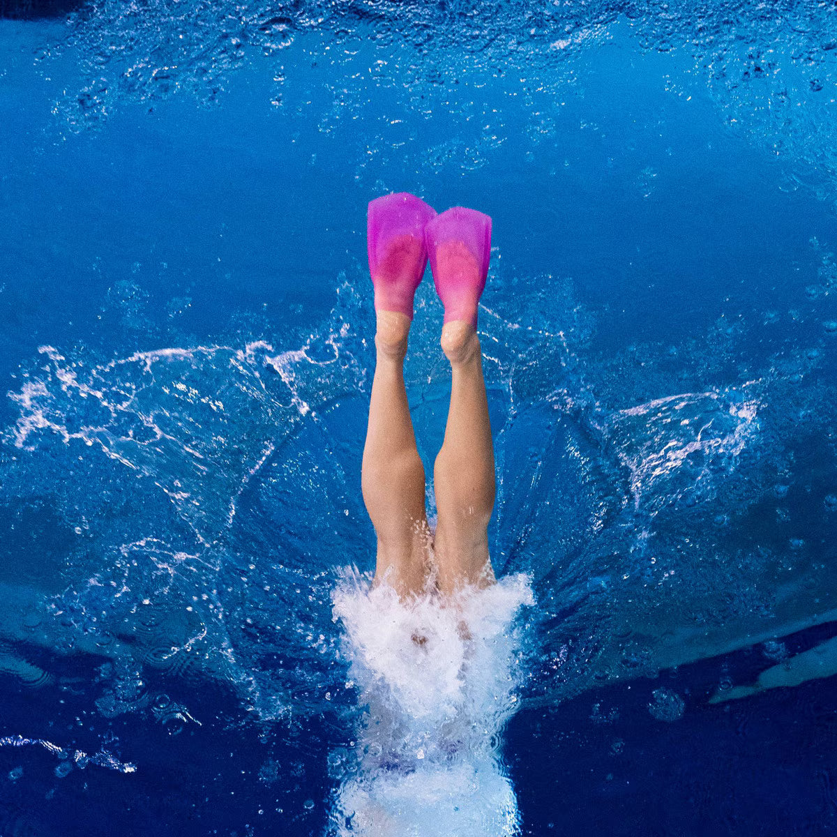 High-angle view of a swimmer diving into blue water, showing legs and translucent deep pink FINIS SlipFin Pro swim fins pointing upward.