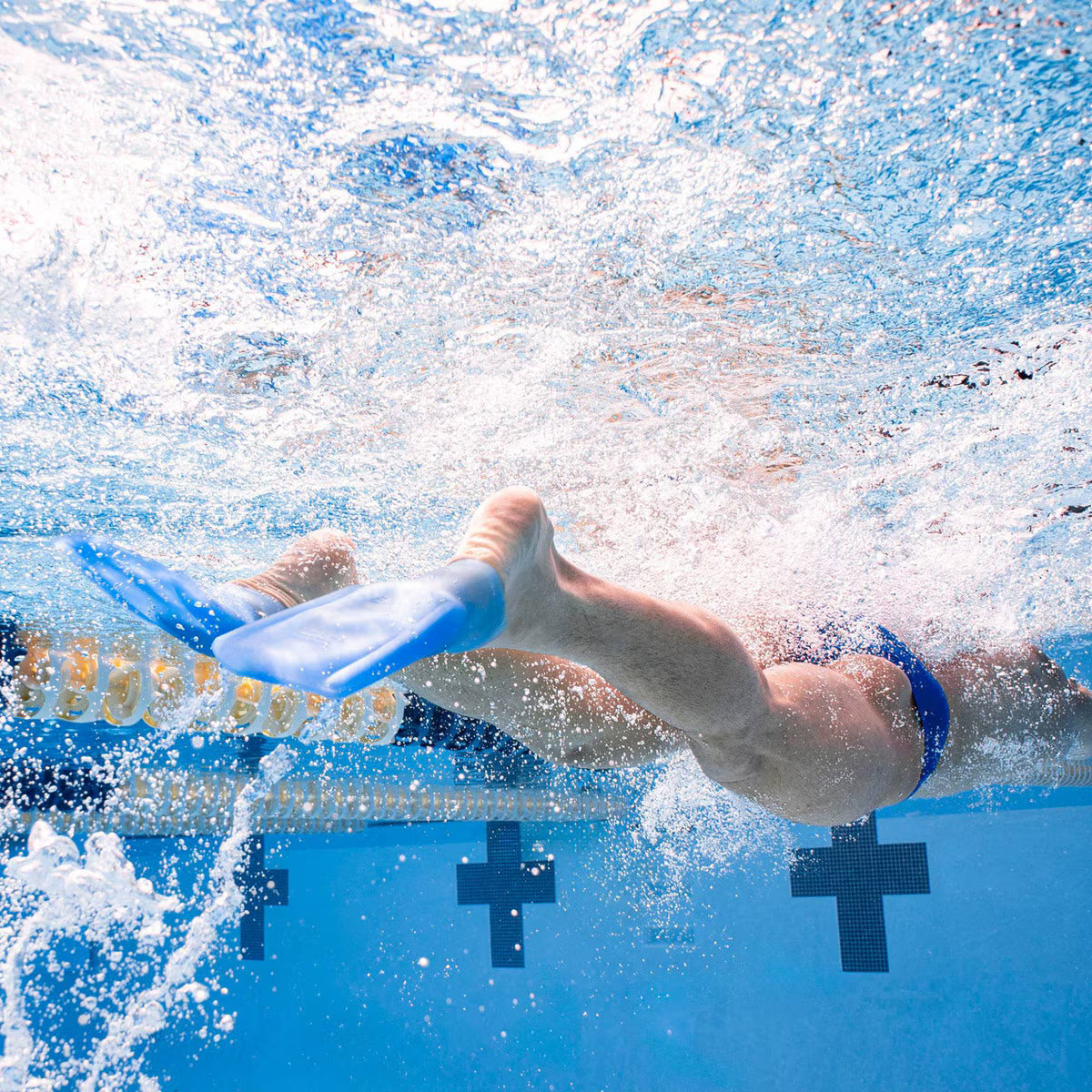 Underwater shot of a swimmer’s legs kicking with translucent blue FINIS SlipFin Pro swim fins, creating bubbles.
