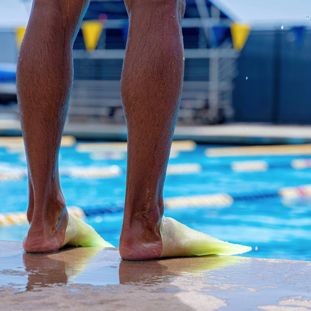 Rear view of a person’s legs standing at the edge of a swimming pool wearing translucent volt green FINIS SlipFin Pro swim fins.